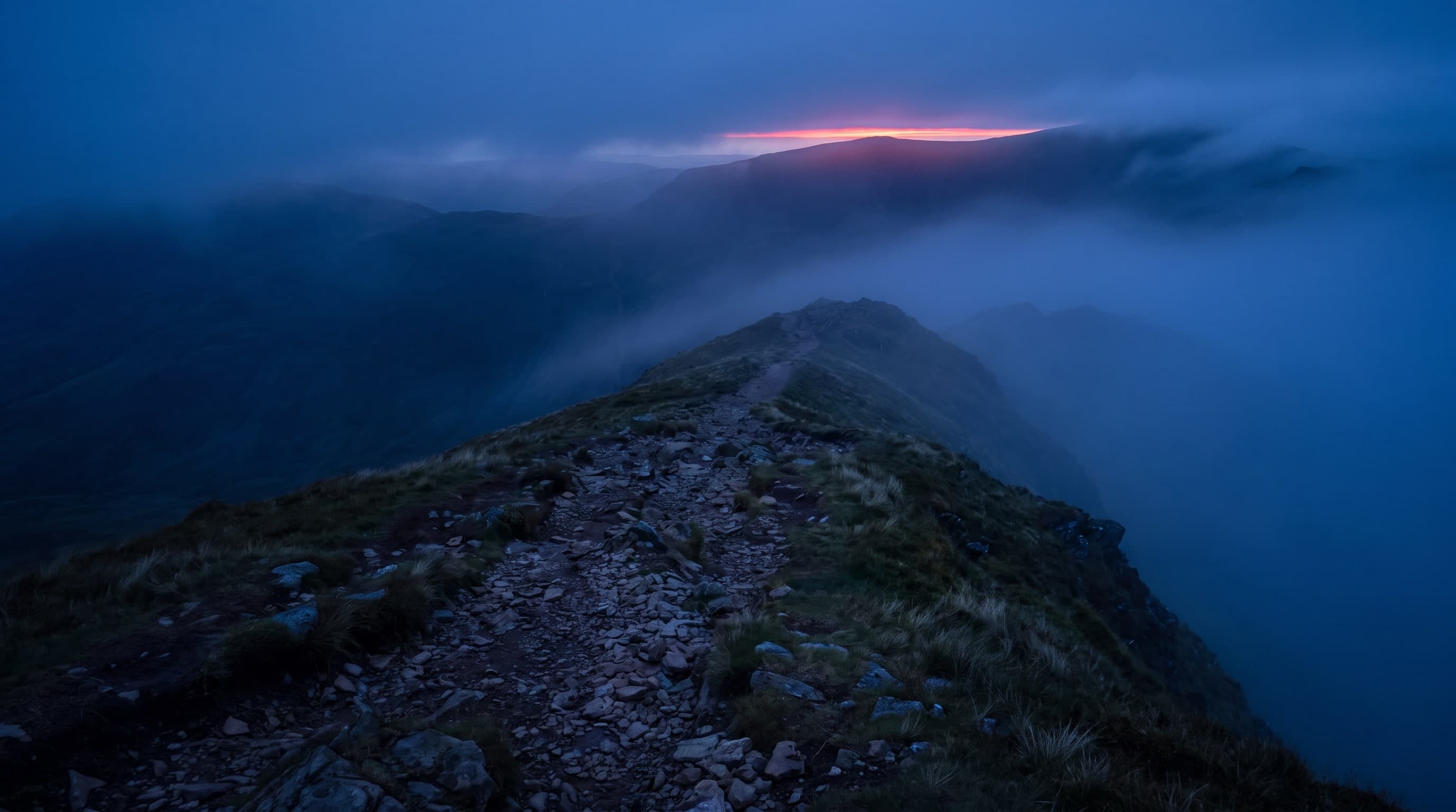 Schmaler Pfad über einen Berggrat im Nebel, am Horizont ein roter Lichtstreif