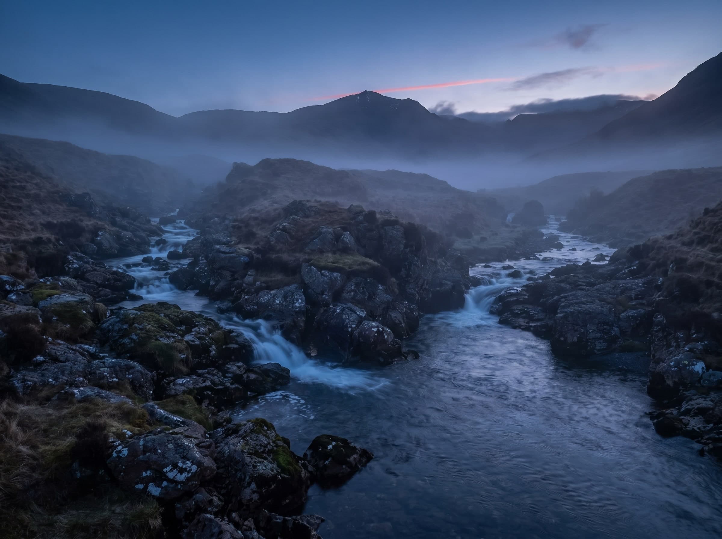 Zwei Gebirgsbäche fließen zwischen Felsen zu einem Fluss zusammen, Nebel in der Dämmerung